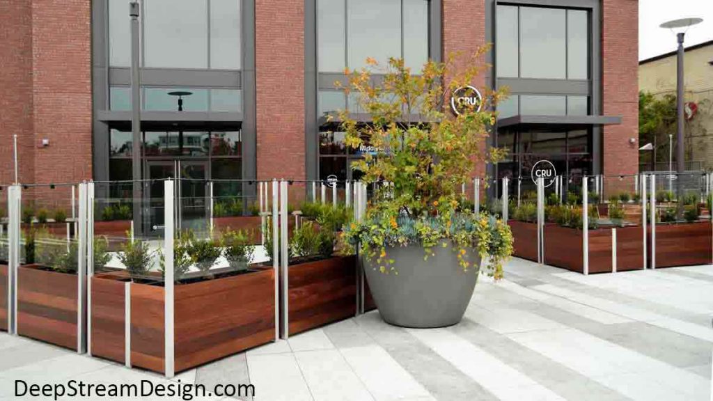 24 large Wood Restaurant Planters planted with bushes create a moveable modular glass screen wall for urban upscale restaurant outdoor seating area in front of a brick building.