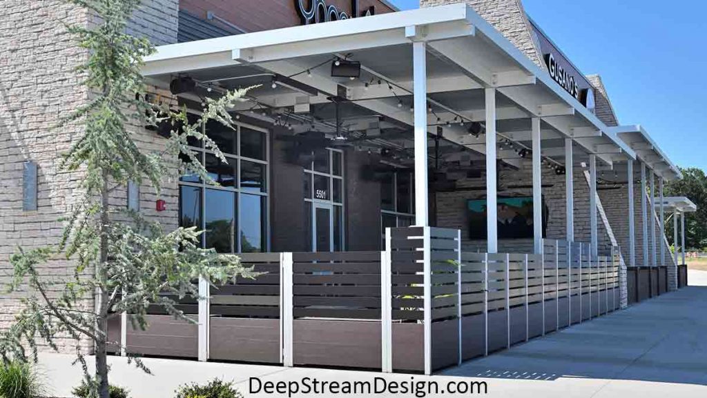 The exterior view of a large Ipe Brown modular Multi-Section Commercial Wood Planter and aluminum screen wall installation, complete with gates, bench seats, and landscaping, that encloses a restaurant’s outdoor smoking area.