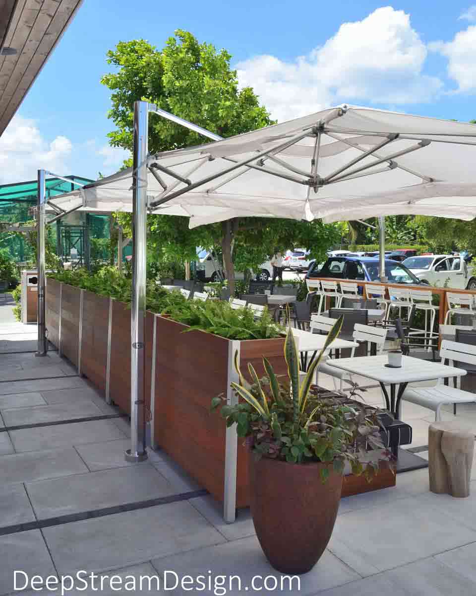 Tall movable rectangular wood Planters on Wheels create a divider between food service and dining areas under large white umbrellas at an upscale outdoor restaurant on a tropical island.