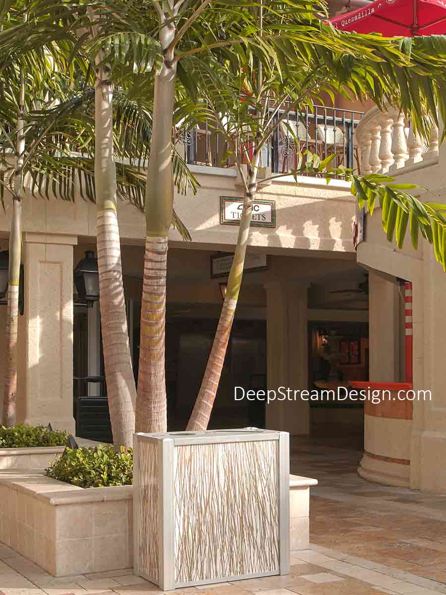 A dual-stream DeepStream Audubon modern commercial combined recycling and trash receptacle with 3form Eco-Resin panels is pictured in the courtyard of an upscale, sophisticated, Mediterranean-style shopping and office complex landscaped with towering palm trees and dark green shrubs.