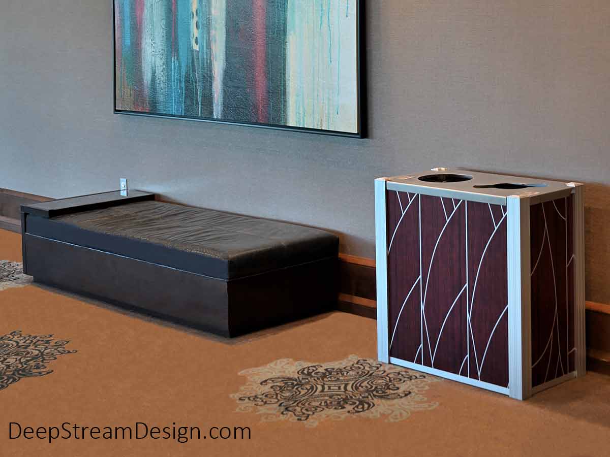 DeepStream Audubon dual-stream modern commercial combined recycling and trash receptacle in the art-filled lobby of a hotels. Although often crafted with wood or 3form panels, this hotel designer has chosen an Audubon bin with a modern silver and dark cherry wood art deco design on an aluminum skin.