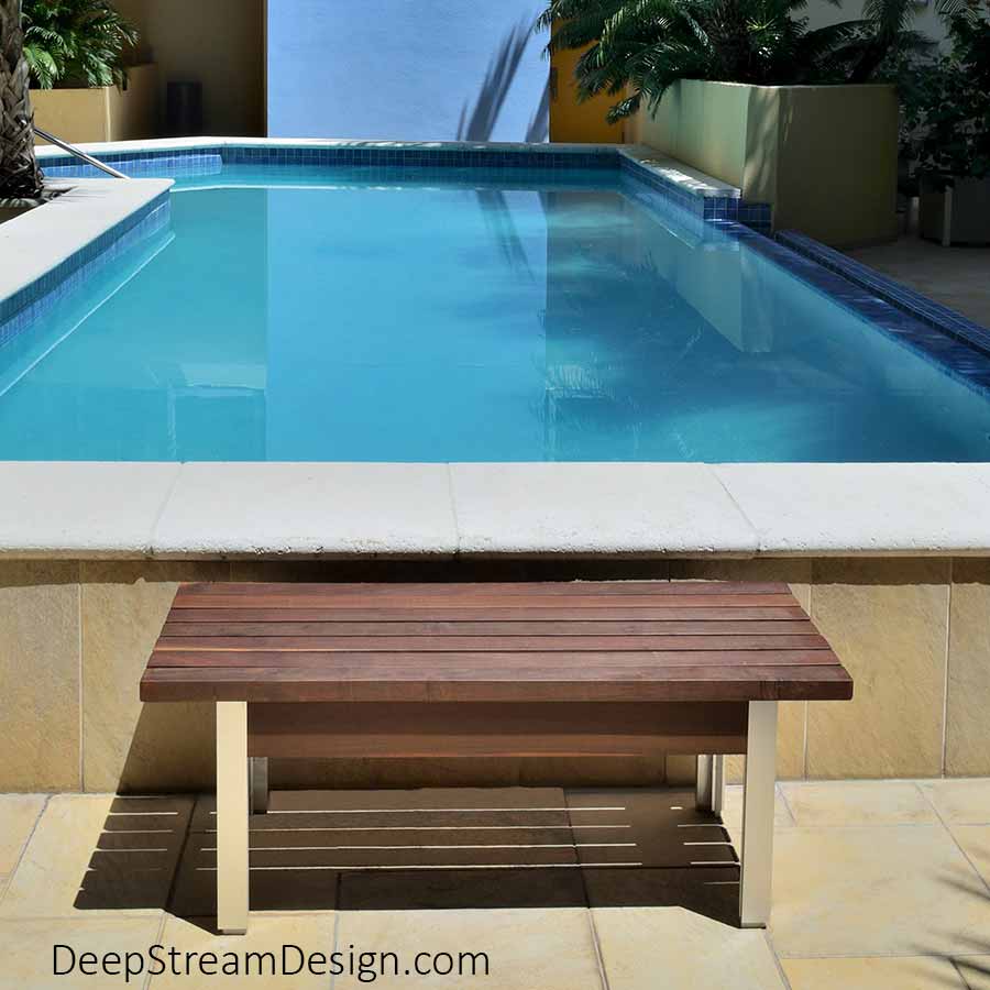 The Perfect Outdoor Bench crafted of dark brown Ipe tropical hardwood and silver anodized aluminum photographed in front of a turquoise blue pool surround by lush dark green tropical plants in the central courtyard of a Miami boutique condominium with blue walls and golden tone natural stone tile.