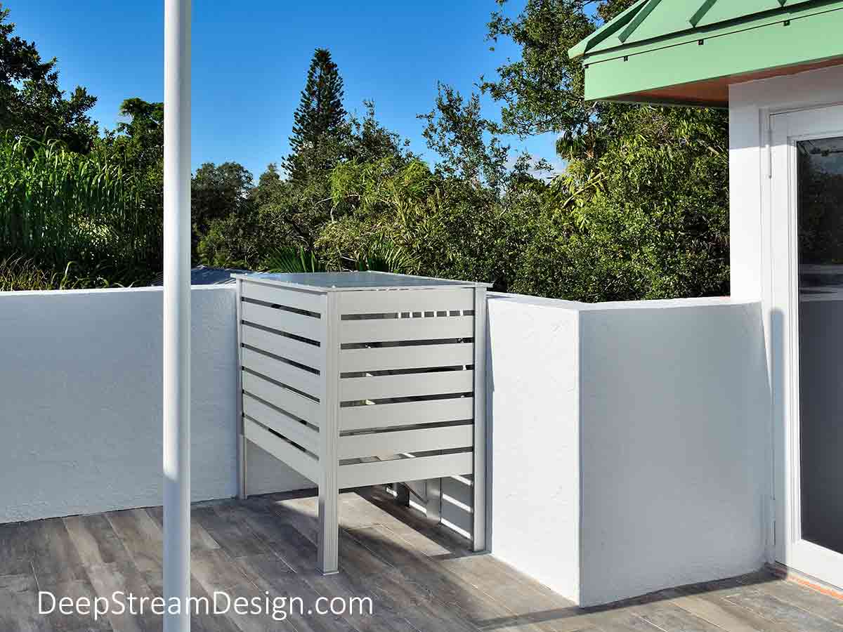 A dangerous electrical utilities panel on a residential roof deck used for sunbathing, BBQ, and dining is covered by a silver aluminum Equipment Screen for Utilities with a flip up lid and removable panels for access.