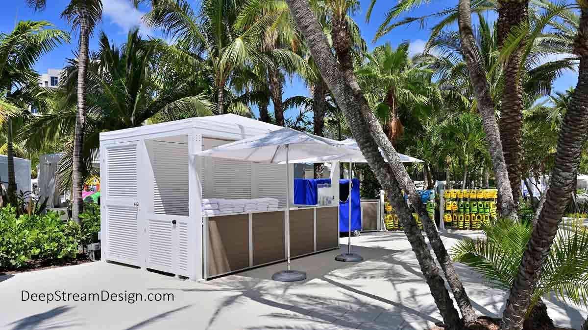 The side view of a large, all-white, outdoor activities center with a canvas top under tall green palm trees with large white umbrellas at a tropical resort waterpark. The Weatherproof Resort Activities Desk and Counter that makes up the entire front of the structure is supported by a series of Weatherproof Cabinets crafted with Aged Hardwood-colored recycled plastic lumber and DeepStream’s proprietary anodized aluminum frames. In addition to the cabinets, there are louvered doors behind the counter for pool towel storage and white towels neatly folded and stacked on 2 of the 4 counter sections. These are important components of DeepStream's outdoor cabinetry and fixtures.